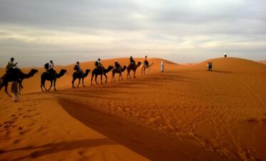 A group traveling on camels over reddish sands under a cloudy sky in a vast desert landscape.