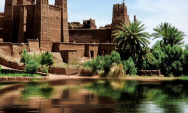 A scenic view of the ancient clay Kasbah Ait Benhaddou by the river in Morocco, under a clear blue sky.